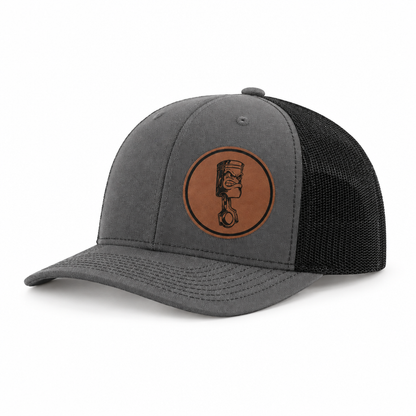 Charcoal gray trucker hat with black mesh back and round angry piston patch on front.