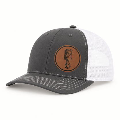 Dark gray trucker hat with white mesh back and round angry piston patch on front.
