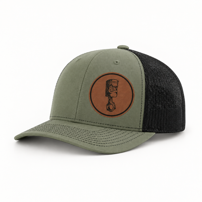 Olive-gray trucker hat with dark mesh back and round angry piston patch on front.