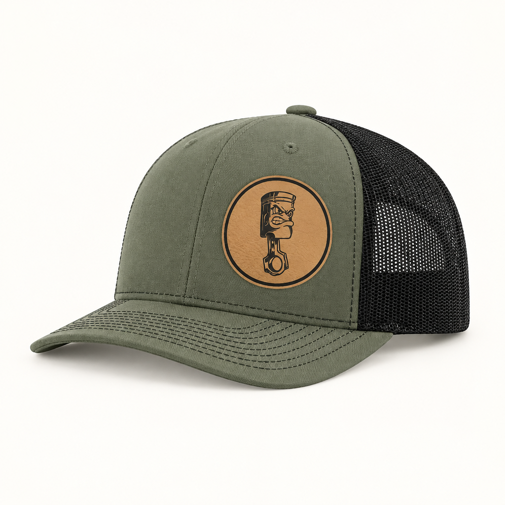 Olive trucker hat with dark mesh back and round angry piston patch on front.