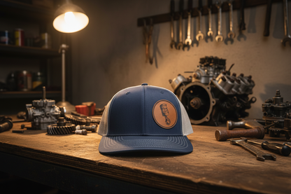 Blue trucker hat with gray mesh back and round angry piston patch