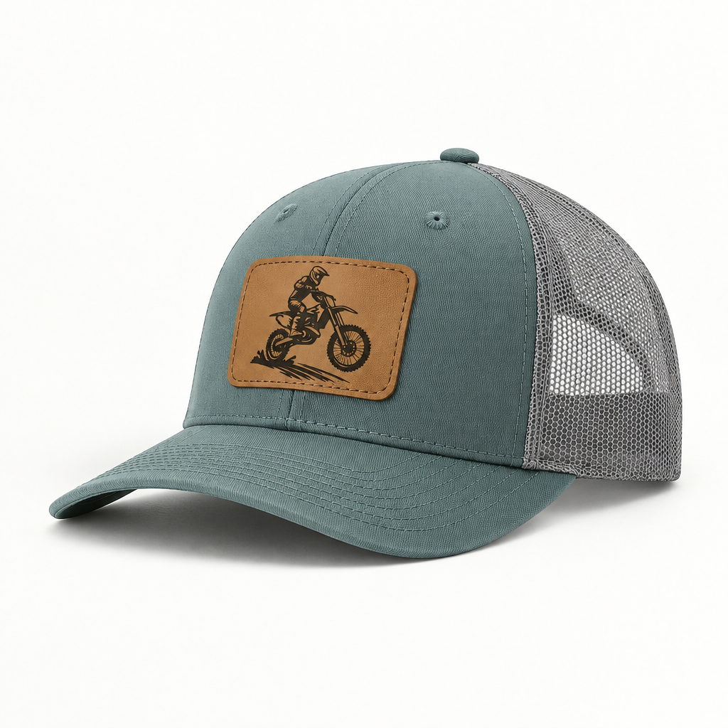 Trucker hat in teal with gray mesh; tan leather patch shows a dirt bike rider.