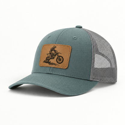 Trucker hat in teal with gray mesh; tan leather patch shows a dirt bike rider.