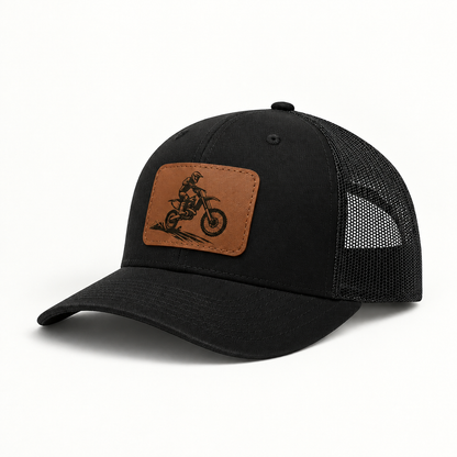 Trucker hat in black with black mesh; brown leather patch shows dirt bike rider.