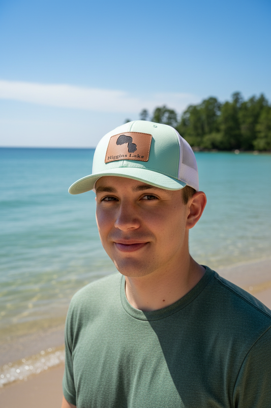 Higgins Lake Michigan leatherette patch trucker hat worn by model on crystal-clear lake shoreline with pine trees – Keeko Designs
